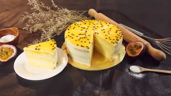 Bánh cheesecake chanh leo đã cắt một miếng, cùng với các dụng cụ làm bánh và trái chanh leo trên bàn.